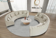 A modern curved sectional sofa in light beige upholstery arranged in a U-shape around a round marble-top coffee table on a large gray area rug, shown in a bright contemporary living room.
